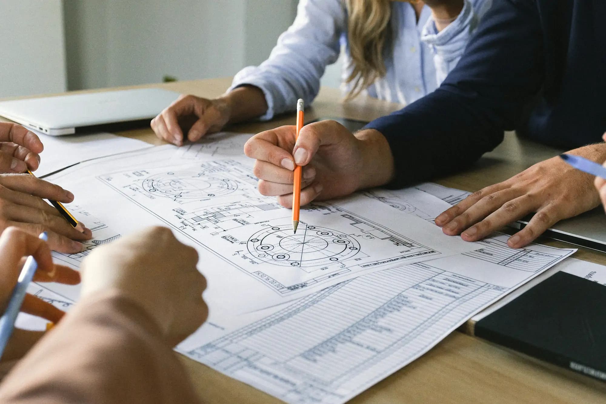 Several people's hands point pencils at technical drawings spread across a wooden table.