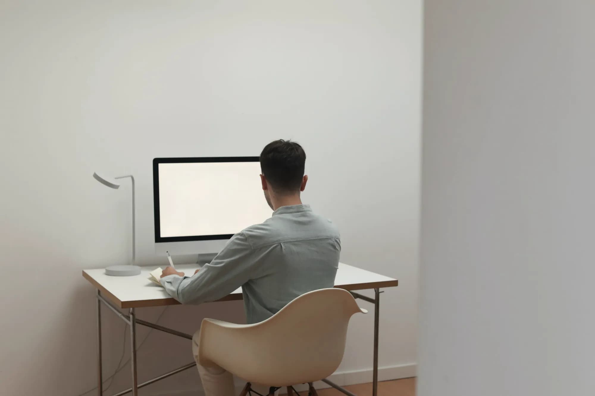 A person seen from behind sits at a white desk in a bright, minimal room, writing in a notebook beside a desktop monitor and a desk lamp.
