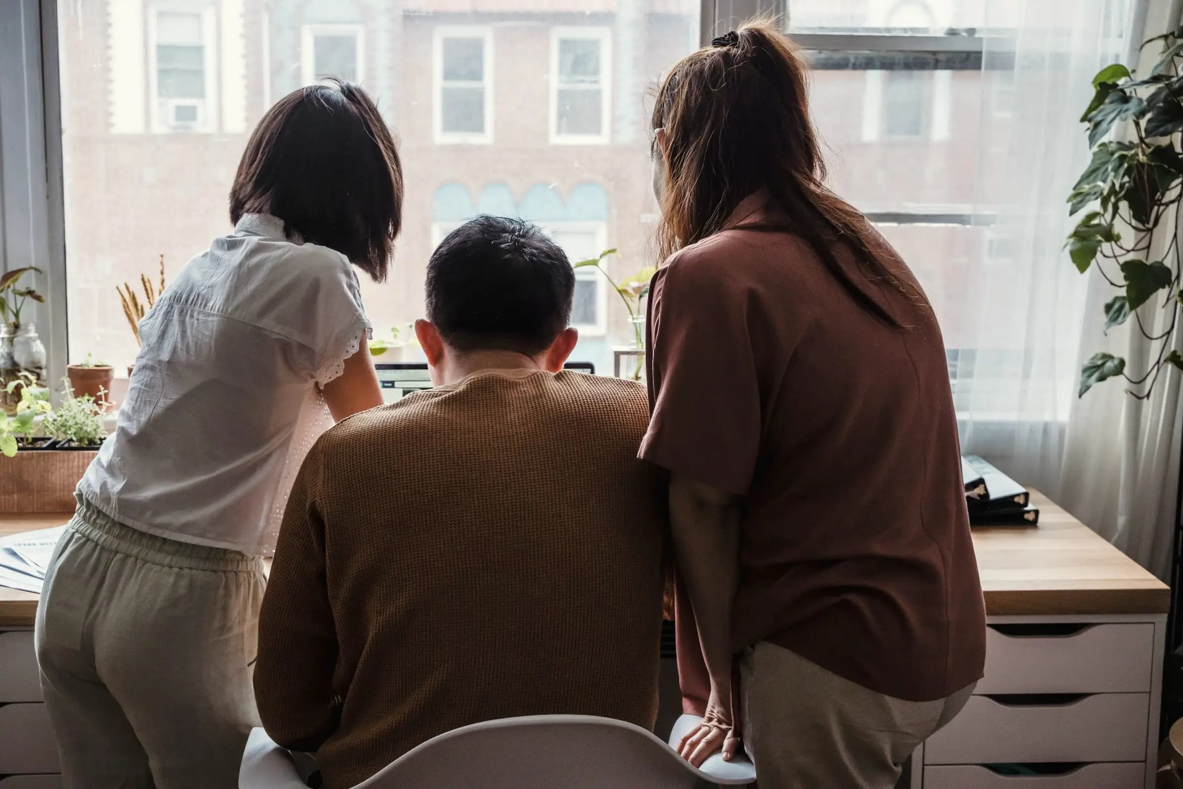 Three people seen from behind gather around a laptop at a desk beside a bright window with houseplants.