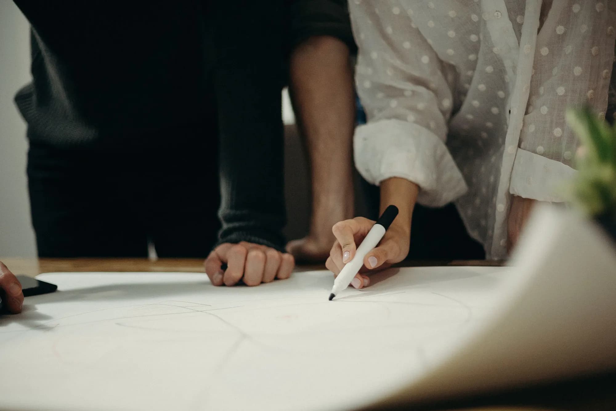 People lean over a table while one of them sketches on a large sheet of paper with a marker.
