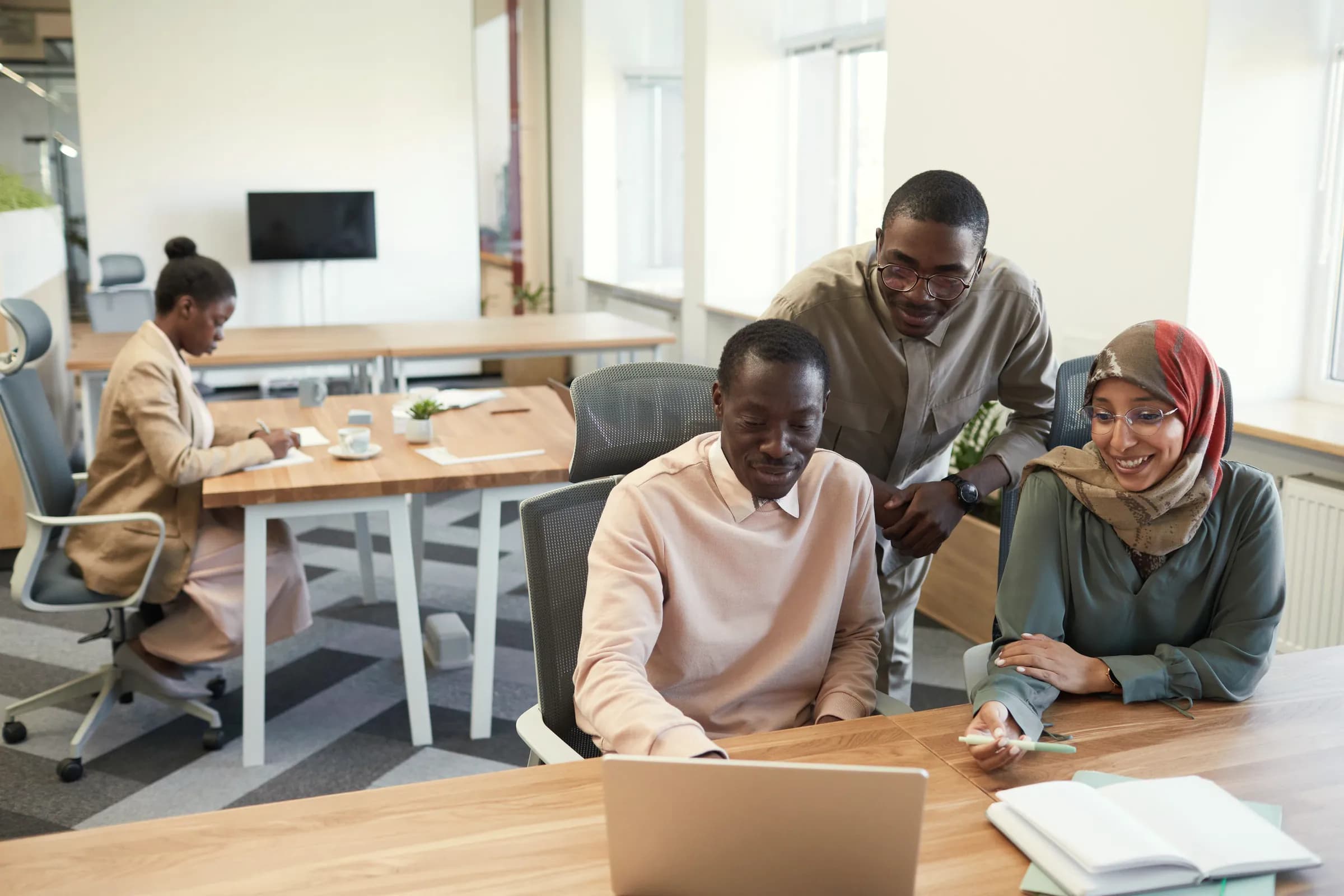 Three colleagues gather around a laptop at a wooden desk in a bright open-plan office, while a fourth works at a table behind them.