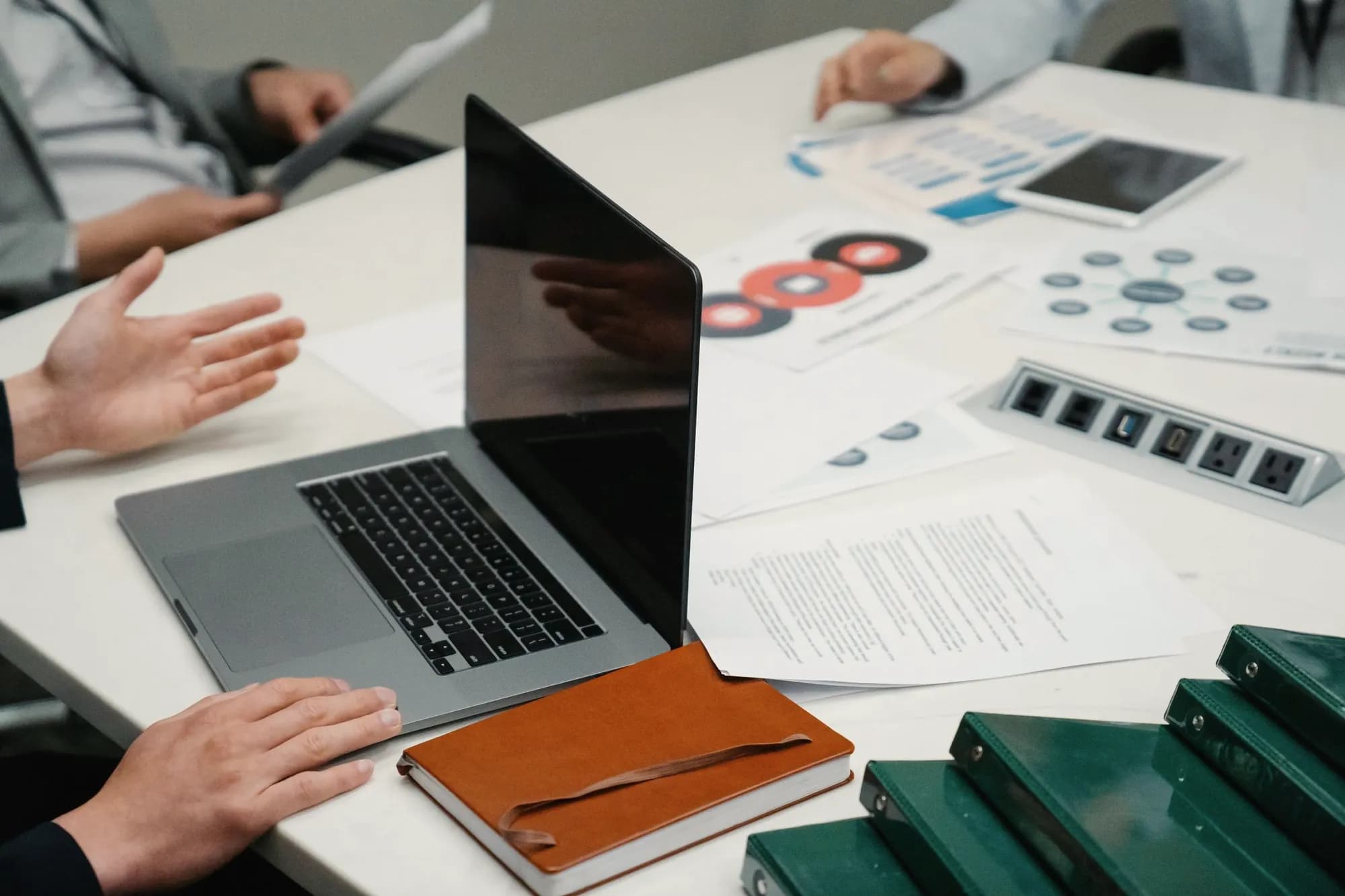 Hands gesture across a meeting table covered with printed reports and diagrams, beside a laptop, a notebook and a stack of green binders.