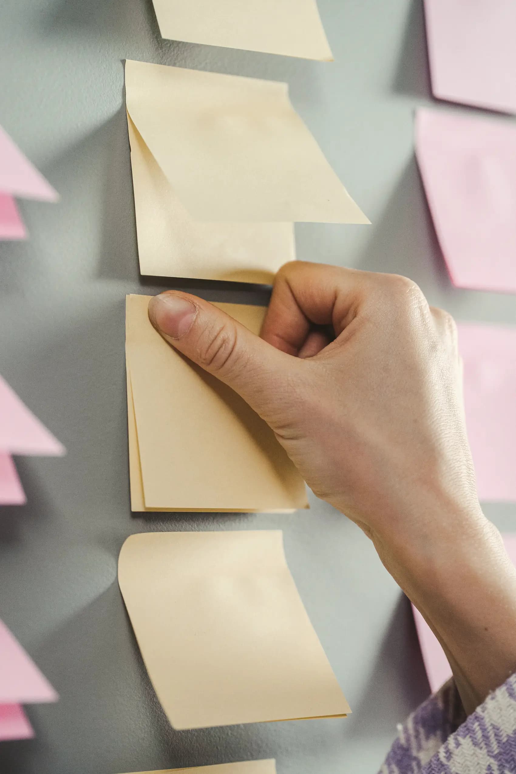 A hand presses a blank sticky note into a column of notes on a grey wall.