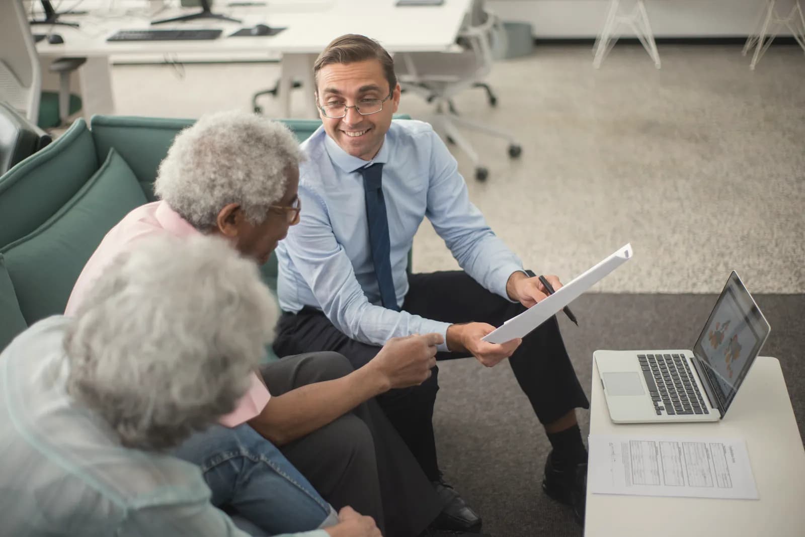 An adviser in a shirt and tie goes through a document with an older couple, a laptop and a printed form on the table beside them.