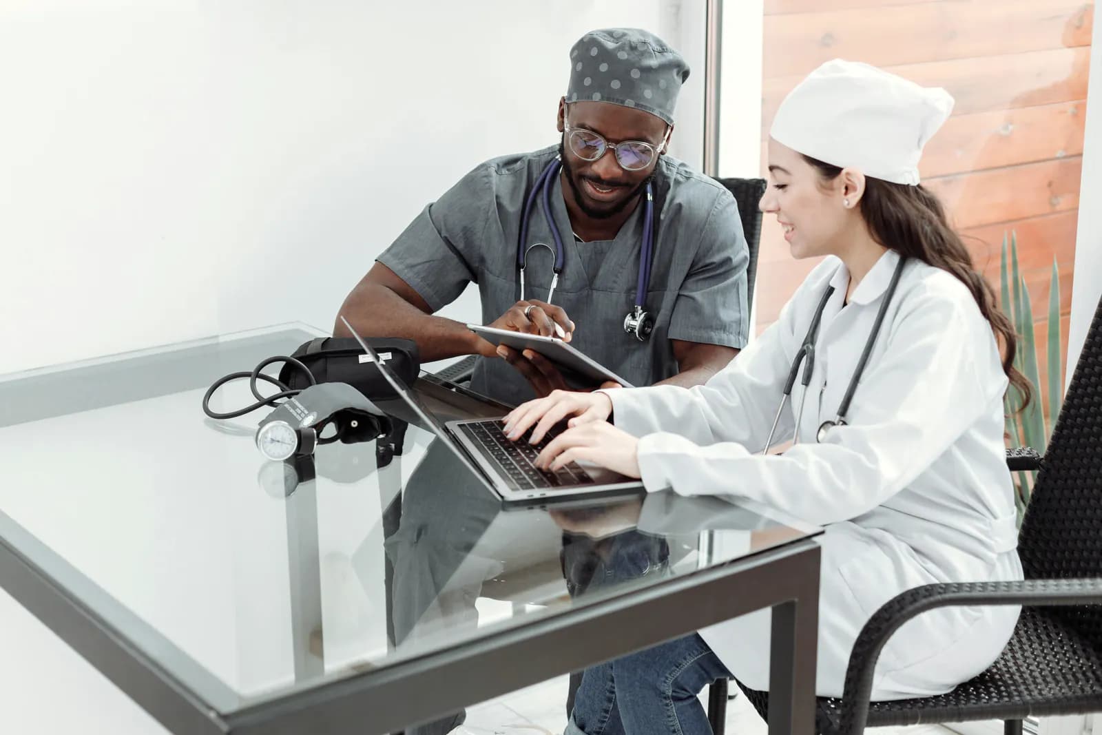 Two clinicians at a glass desk, one holding a tablet and the other typing on a laptop, a blood pressure monitor beside them.