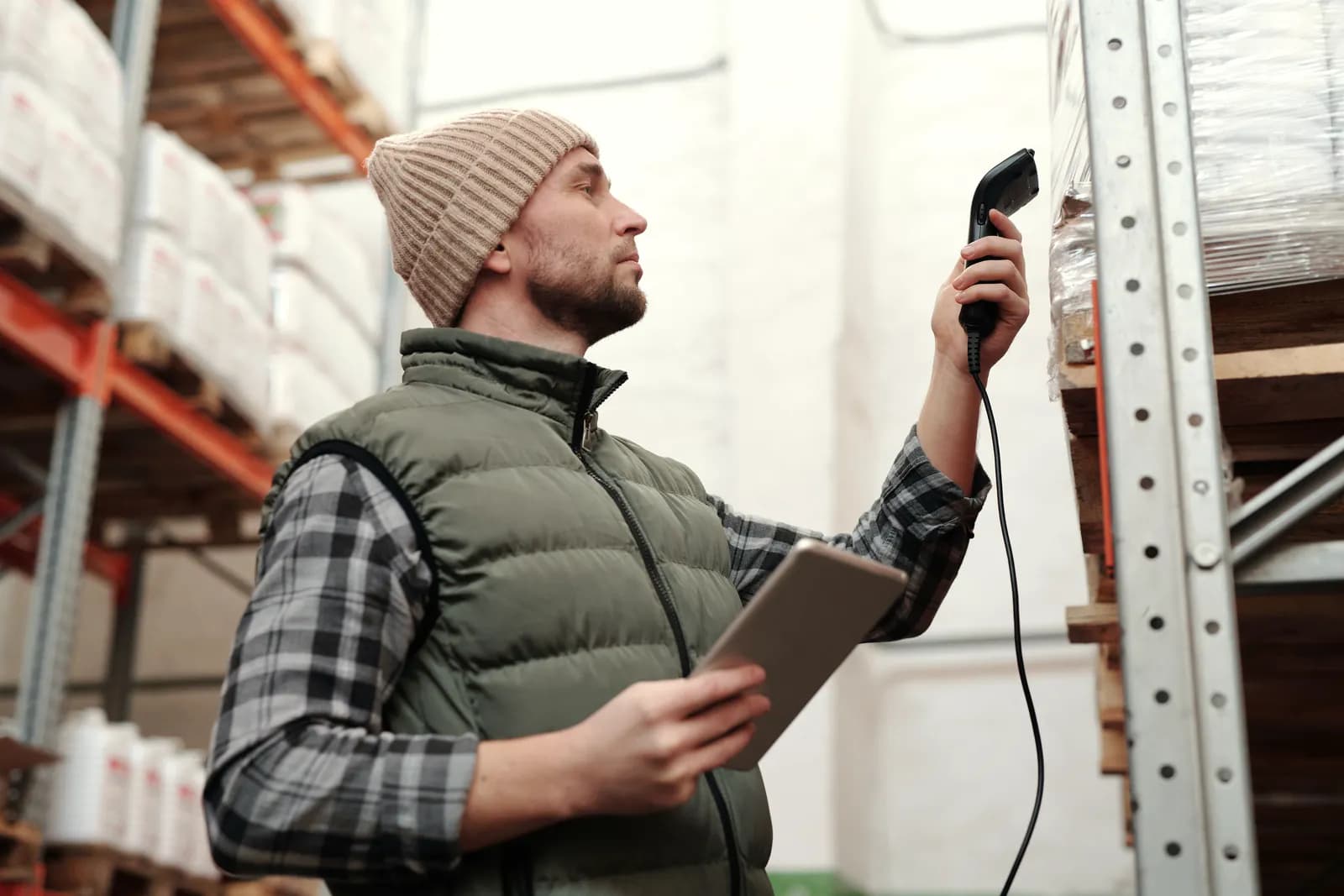 A warehouse worker holding a tablet scans a pallet on a high shelf with a handheld barcode scanner.