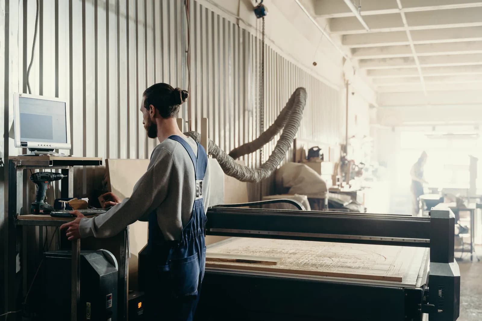 A worker in overalls sets up a large CNC router from its control station in a bright workshop.