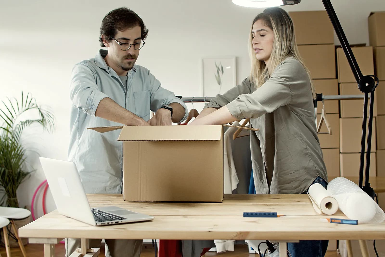 Two people pack an order into a cardboard box at a wooden table, with a laptop, packing paper and a clothes rail around them.
