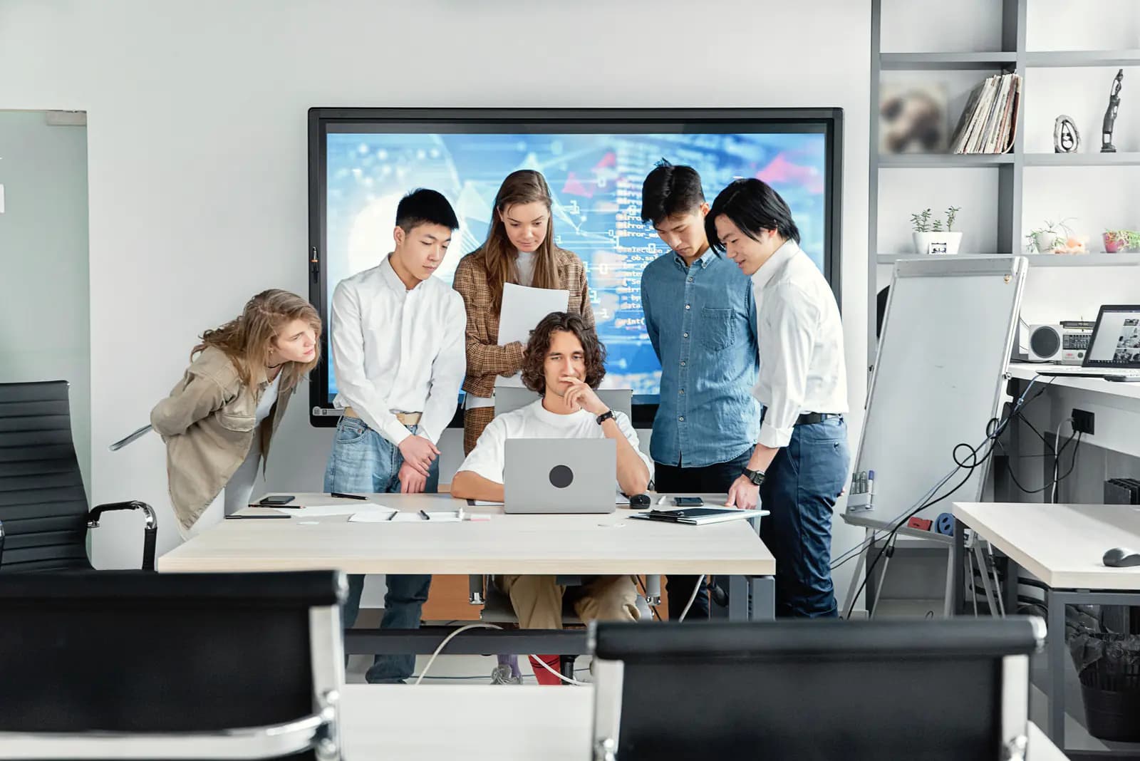 Five colleagues gather behind a seated colleague to look at his laptop, with a large screen on the wall behind them.
