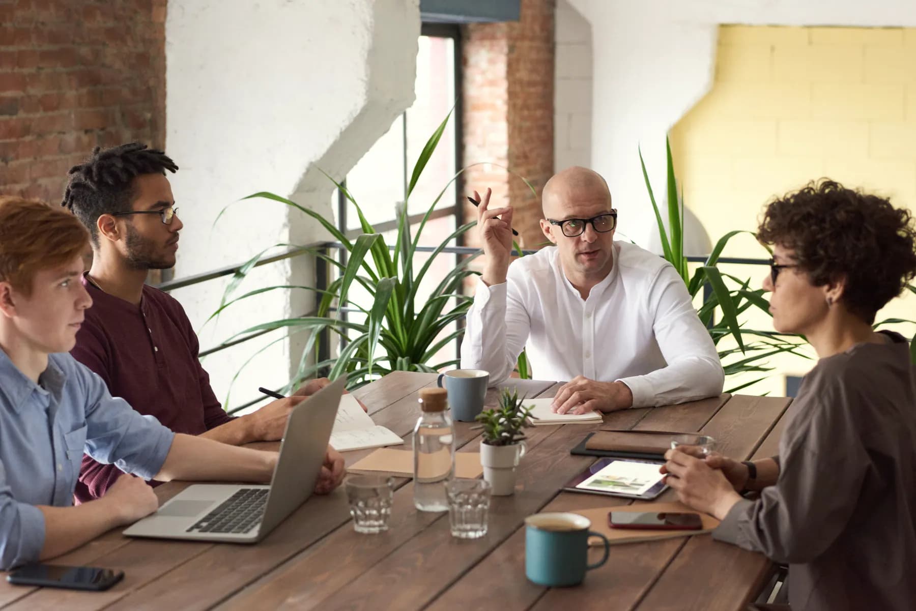 Four colleagues talk around a wooden table with a laptop, notebooks and coffee cups, plants behind them.