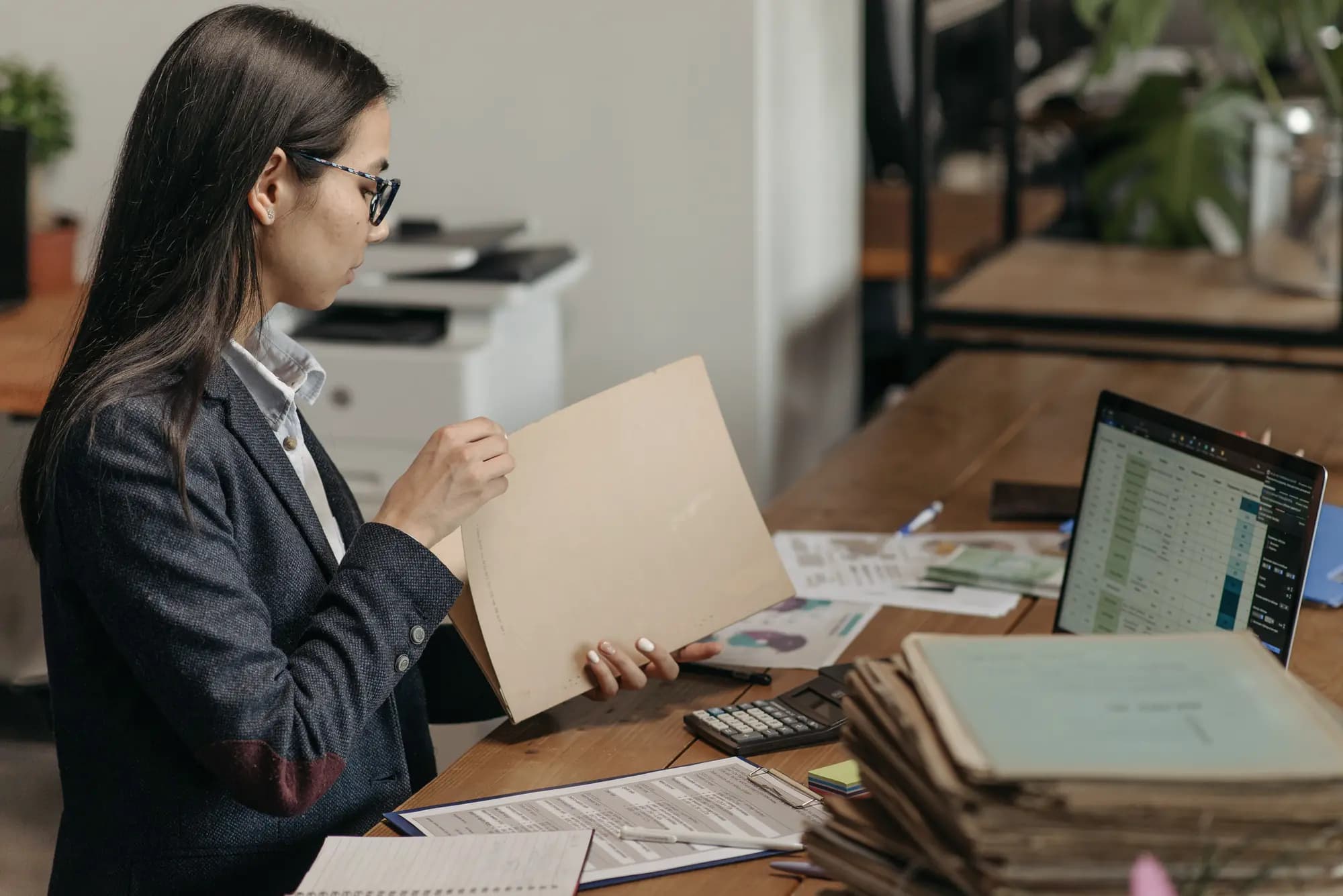 A woman at an office desk works through a folder, with a spreadsheet open on her laptop, a calculator and a tall stack of paper files beside her.
