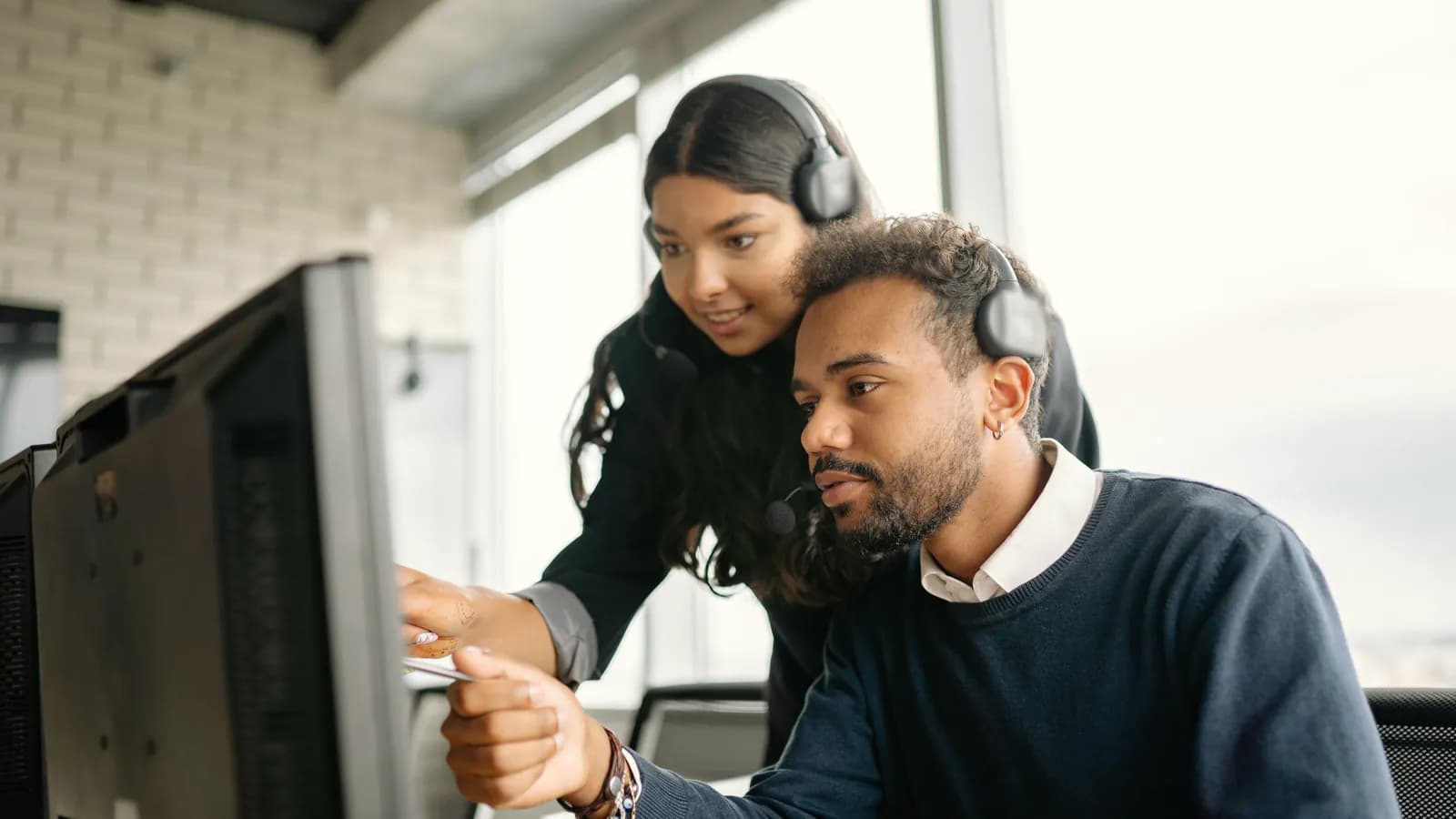 Two support engineers in headsets look at a monitor together, one pointing at the screen.