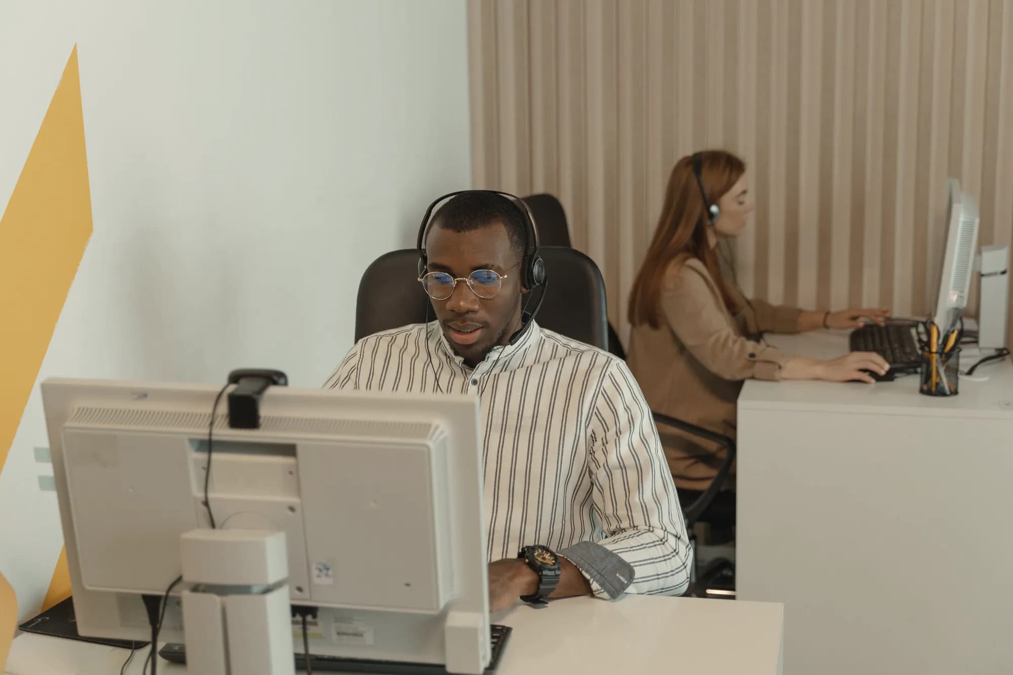 A support engineer in a headset works at a desktop monitor, with a colleague in a headset at another desk behind him.