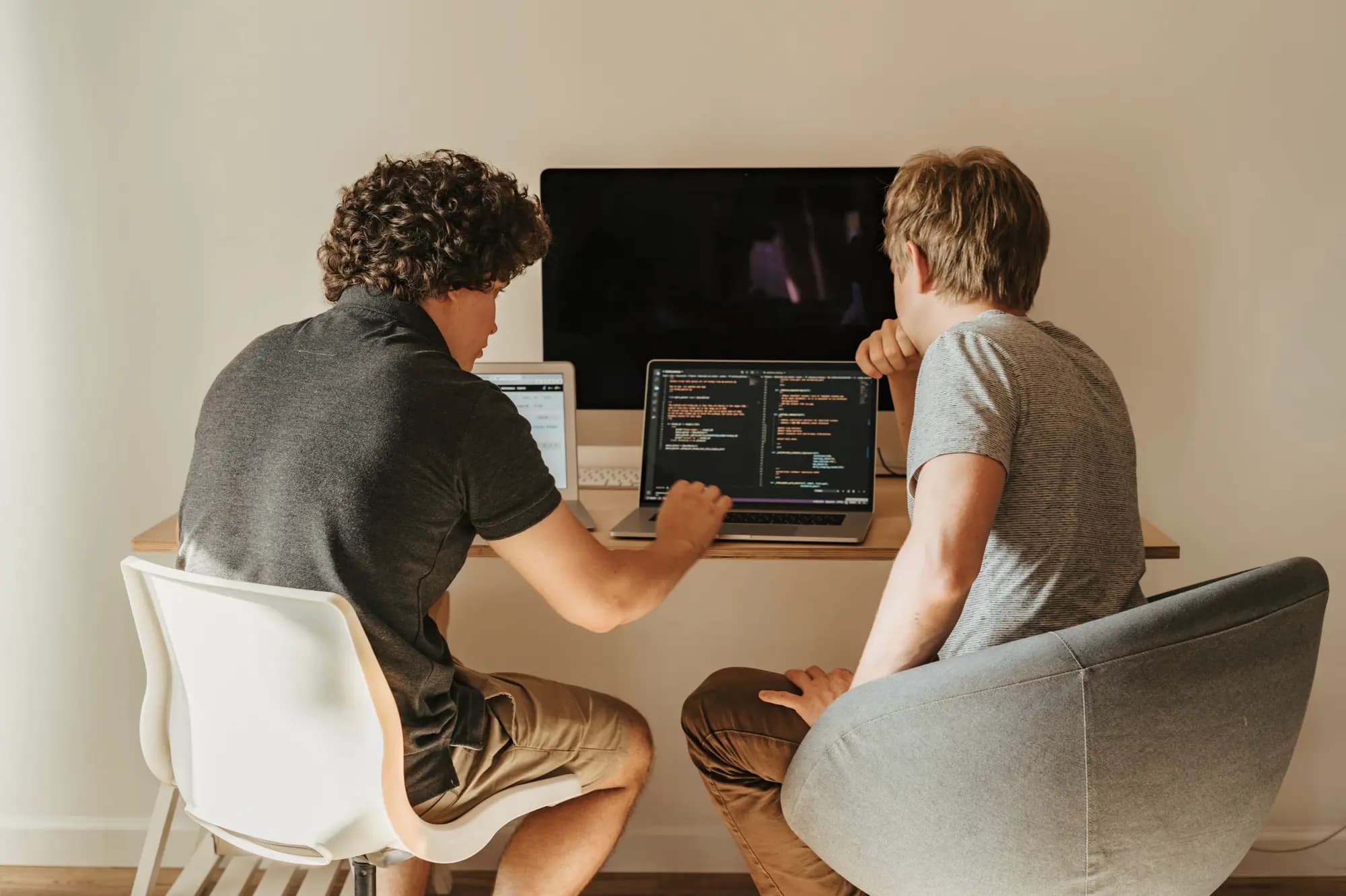 Seen from behind, two developers at a desk review code on a laptop, a large monitor behind it.