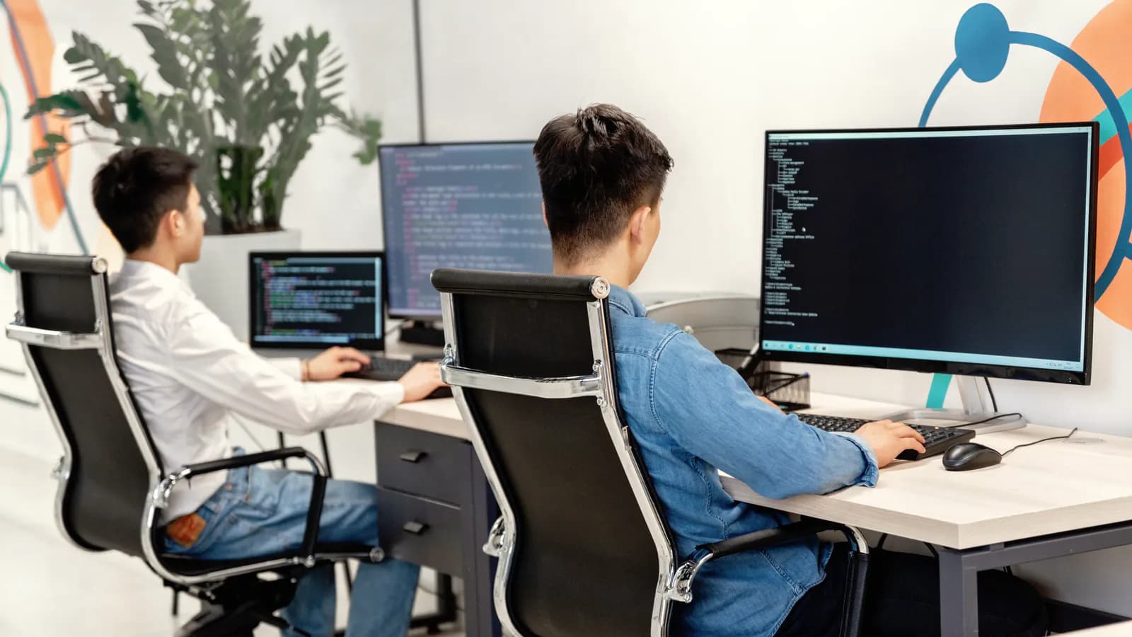 Seen from behind, two developers work at large monitors showing code in a bright office.