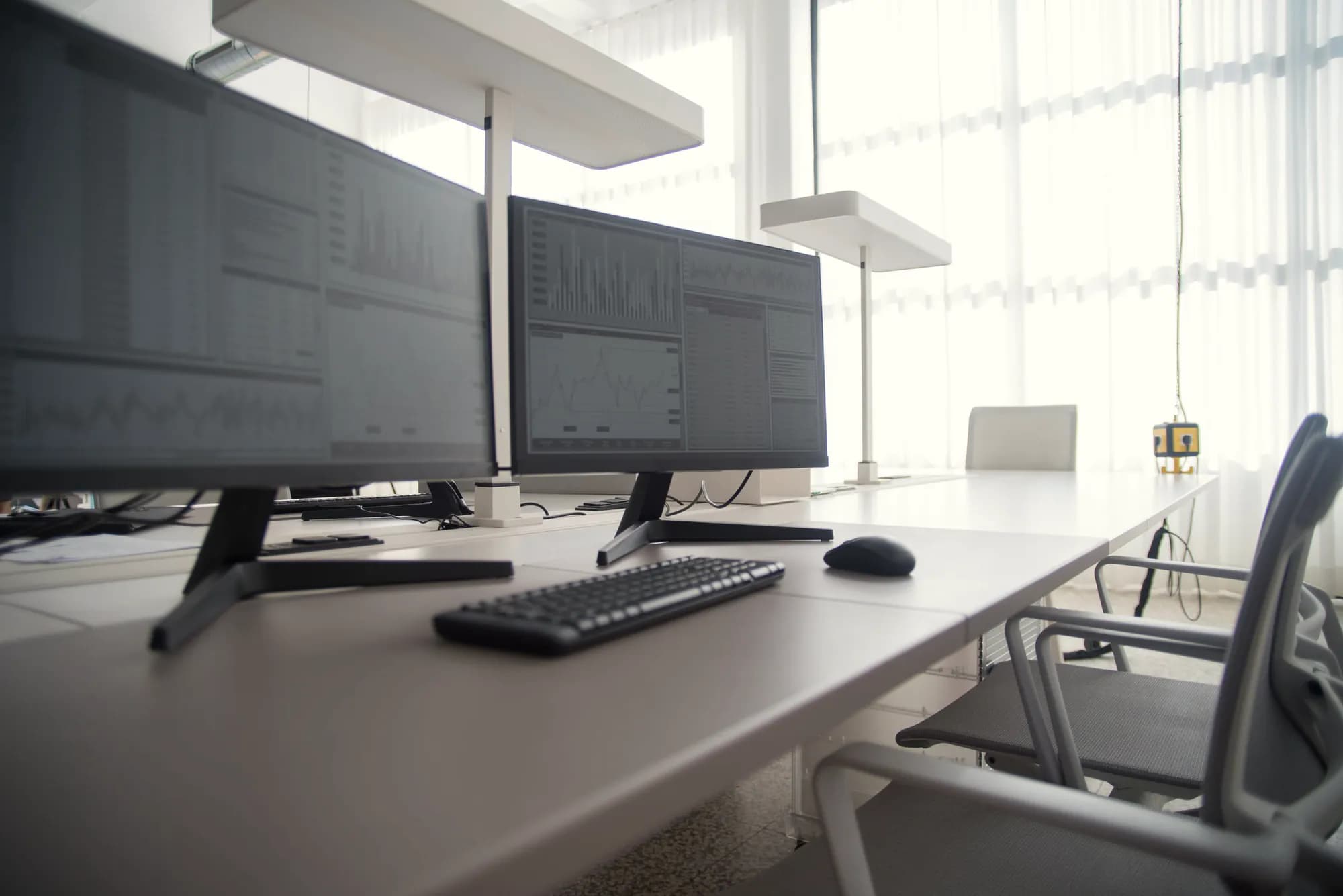 Two monitors showing charts and tables stand on an empty office desk beside a bright window.