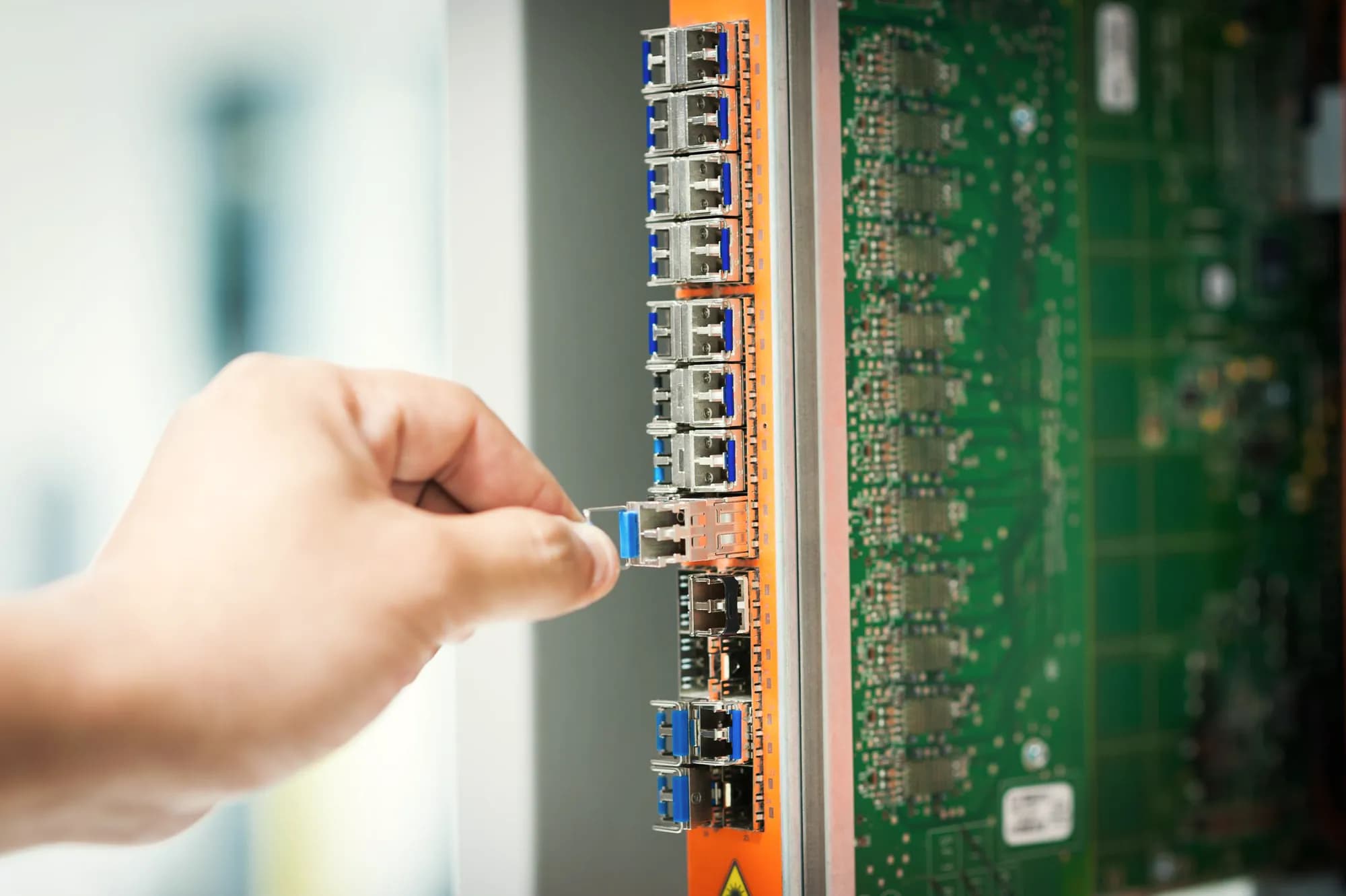 A hand slides a small optical transceiver out of a row of ports on a network switch card.