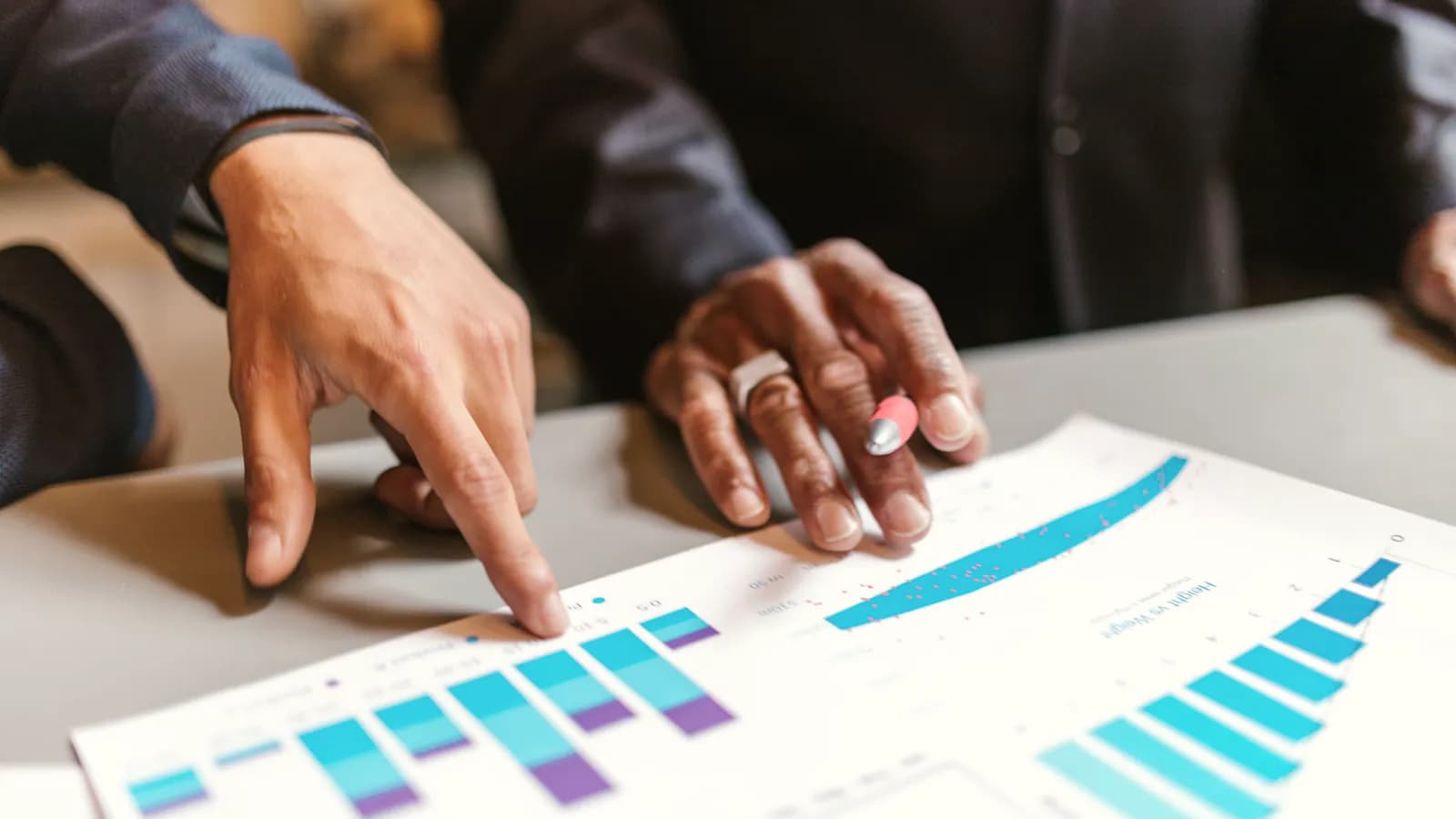 Two people's hands over a printed bar chart, one pointing at a bar and the other holding a pen.