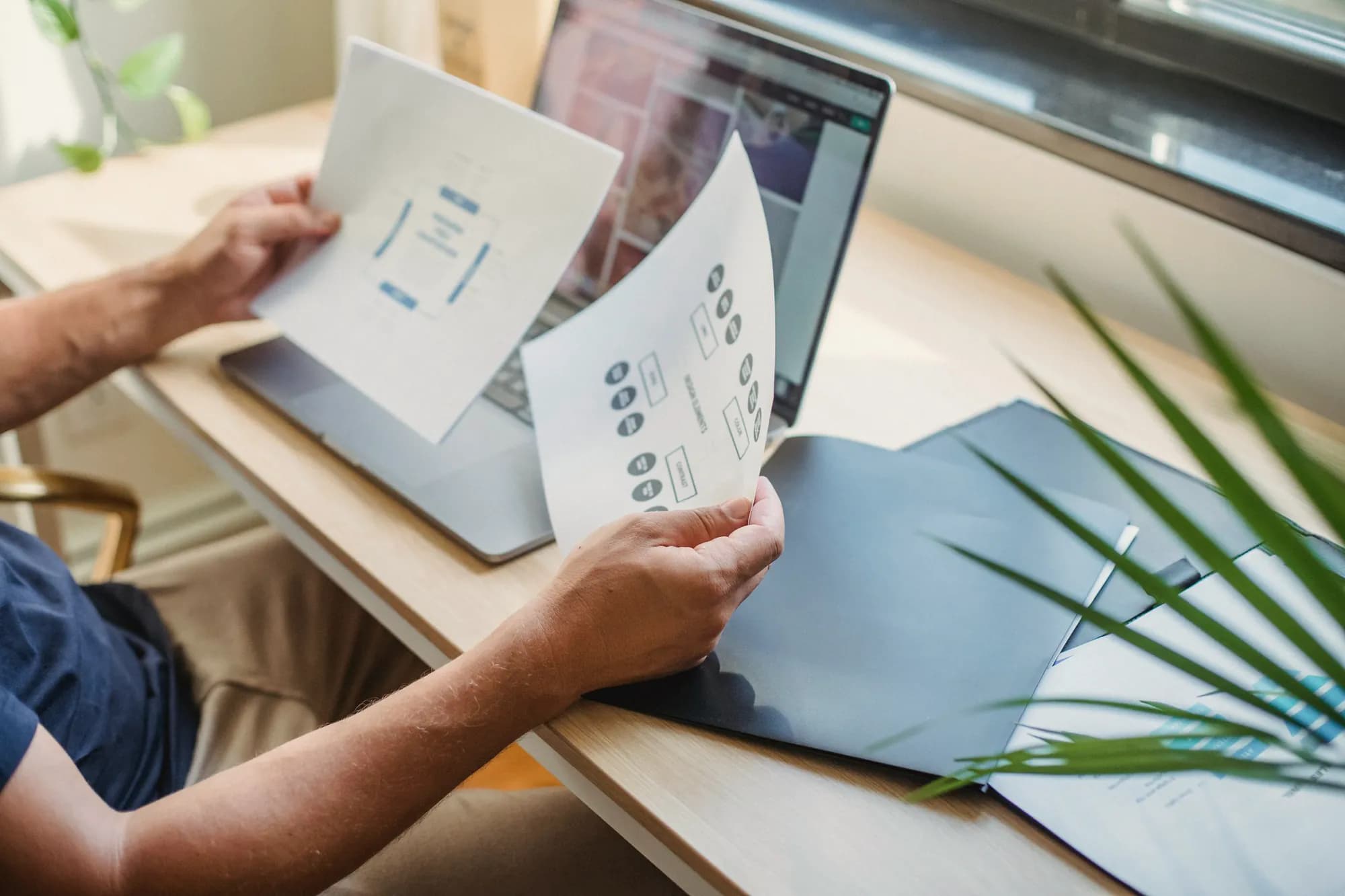 Hands hold two printed pages of diagrams up in front of an open laptop on a desk.