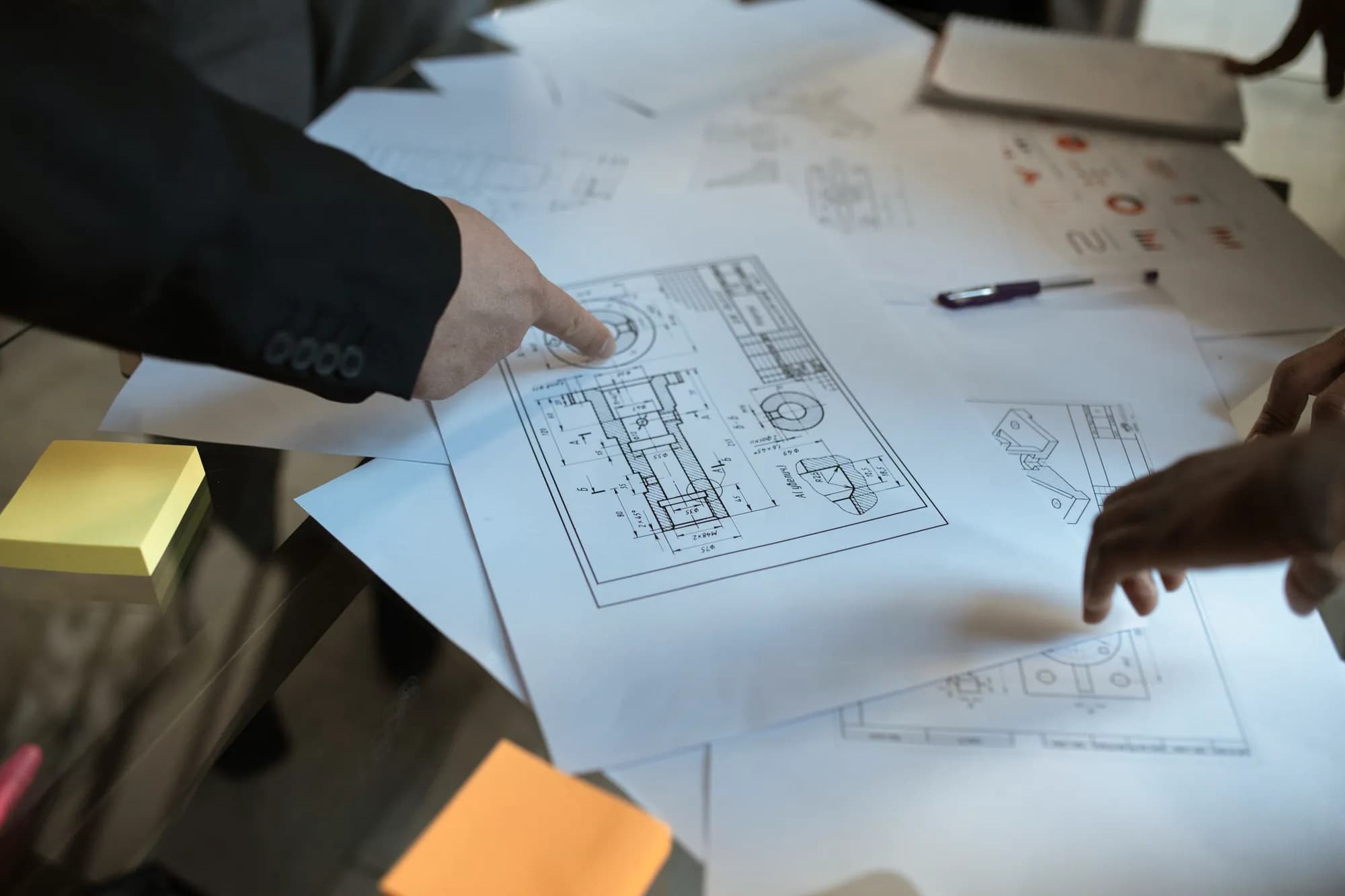 Two people's hands point at technical drawings spread across a table.