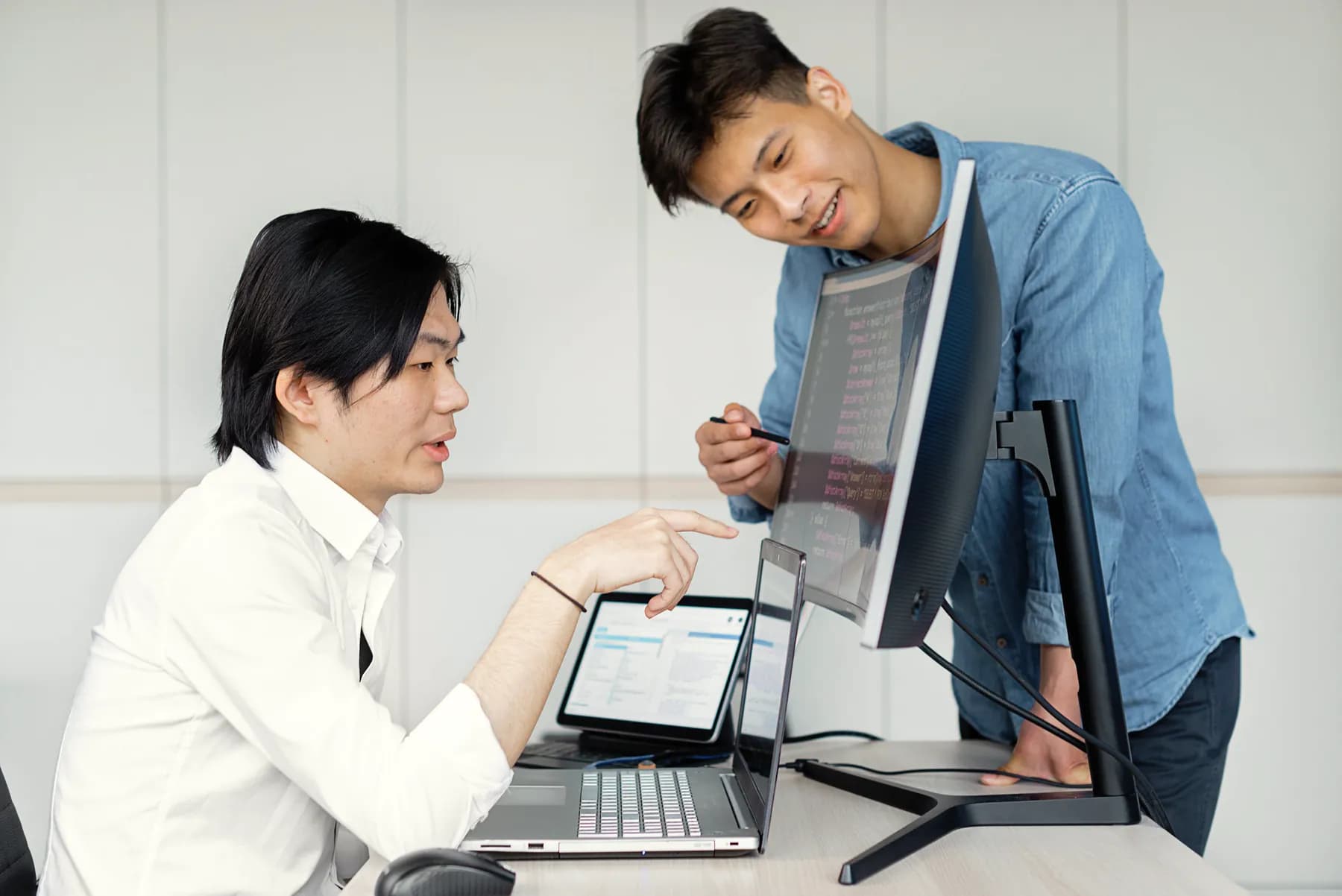 A seated engineer points at his laptop while a colleague in a denim shirt leans over a monitor of code beside him, pen in hand.