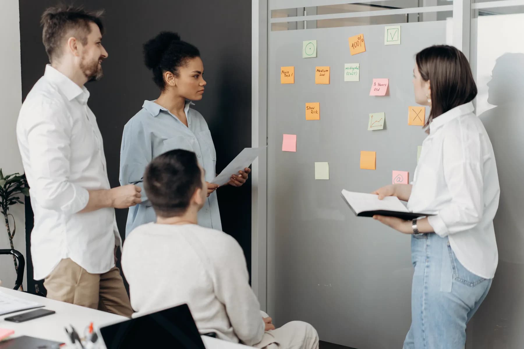 Four colleagues plan together at a wall of sticky notes in a bright office, one of them holding an open notebook.