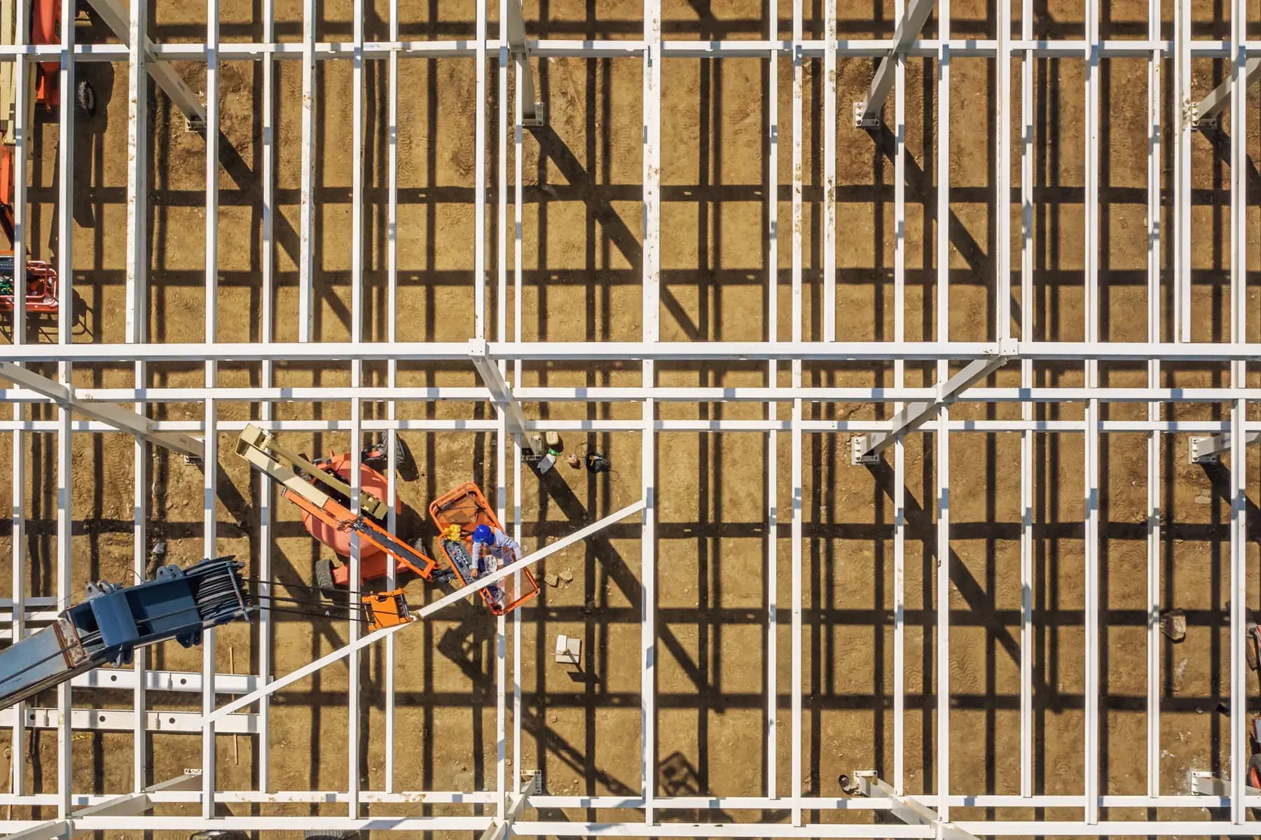 Looking straight down on the white steel grid of a building frame under construction, its shadows falling on bare earth, with a boom lift at one corner.