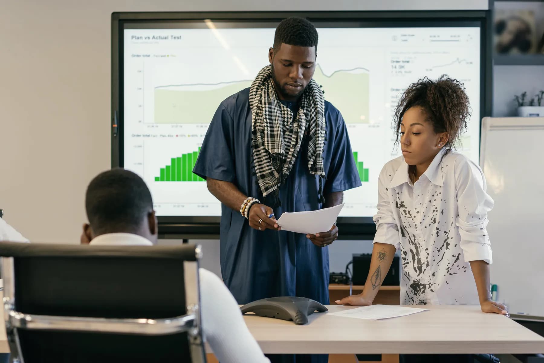 Two colleagues read a printout together in a meeting room, in front of a large screen of charts comparing plan and actual figures.