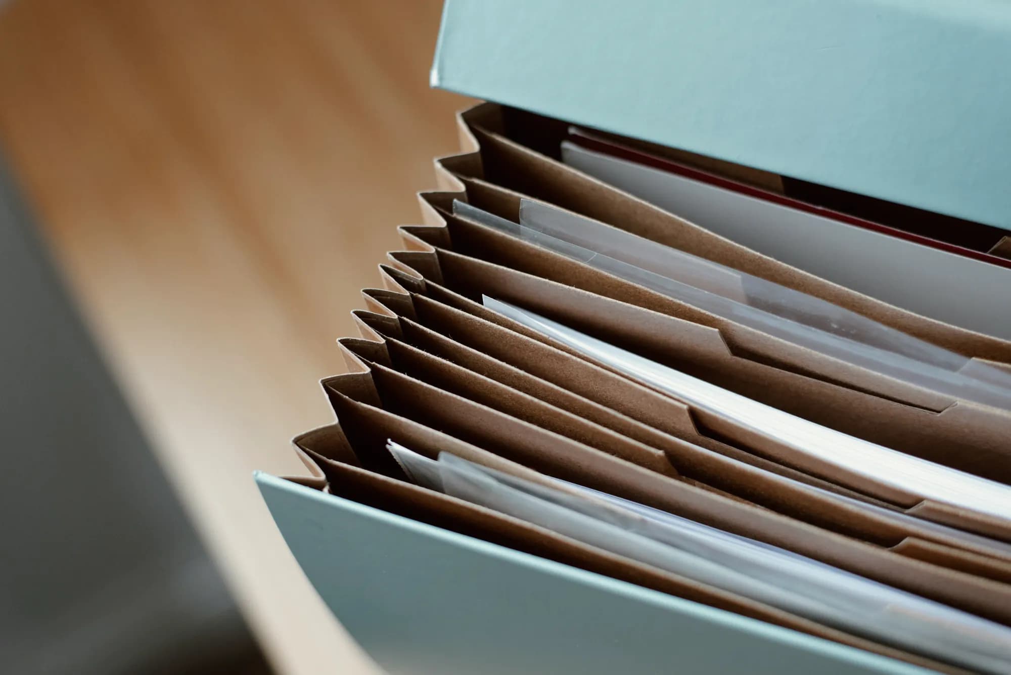 Close-up of an open pale blue expanding file on a wooden desk, its kraft-paper pockets filled with loose sheets.