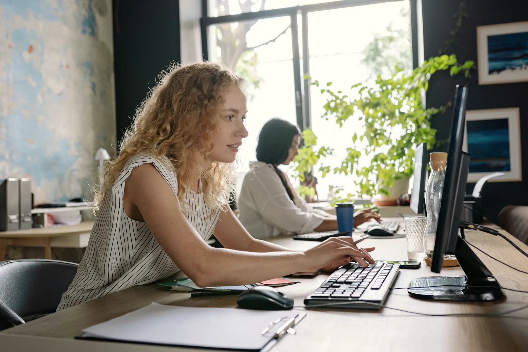 A woman with curly hair types at a desktop computer in a bright office full of plants, while a colleague works at the next desk.