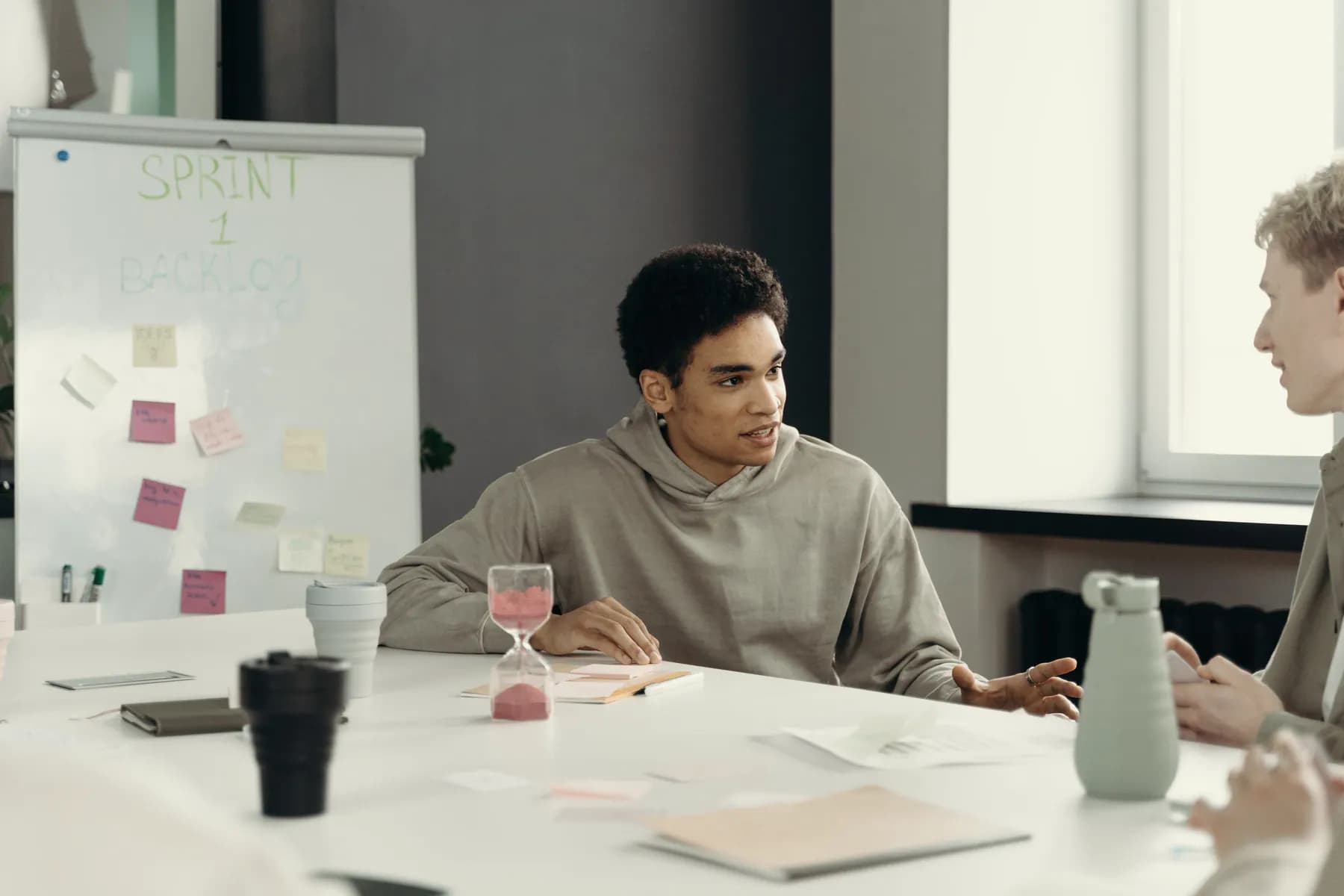 At a meeting table, a young man in a hoodie talks through a plan, a sand timer in front of him and a flip chart of sticky notes headed Sprint 1 backlog behind him.