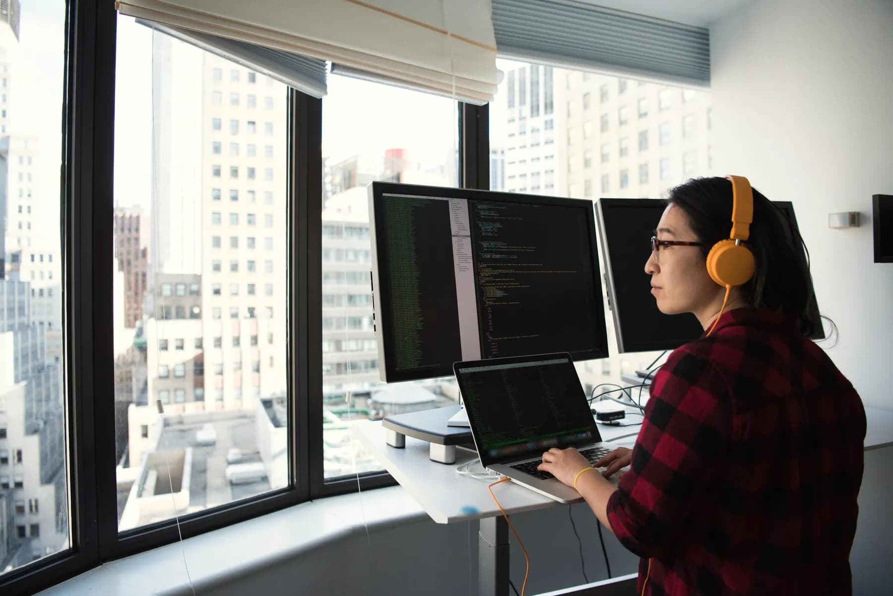 A developer in orange headphones works at a standing desk by a tall window, with code open on two monitors and a laptop.
