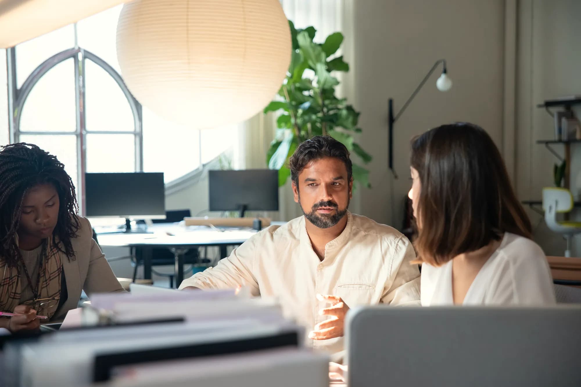Three colleagues talk across a desk in a sunlit office with an arched window and a paper lantern overhead.