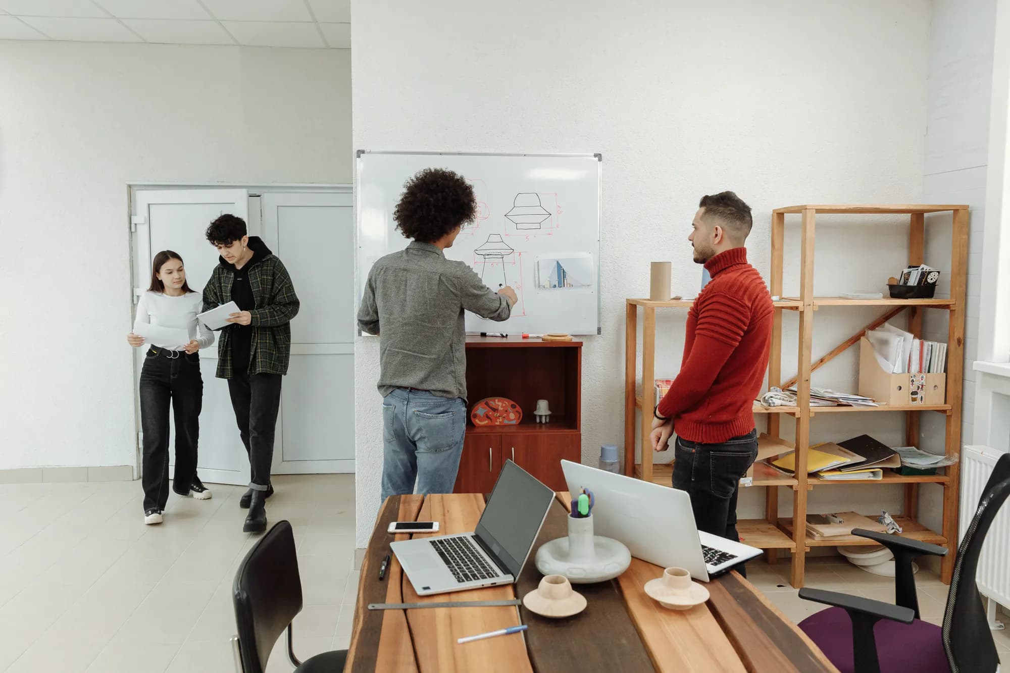 In a bright studio, one colleague sketches on a whiteboard while another looks on and two more walk in reading papers.